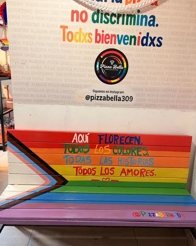 Banca arcoíris de adentro que dice «Aquí florecen todos los colores, todas las historias, todos los amores», bajo el muro del local