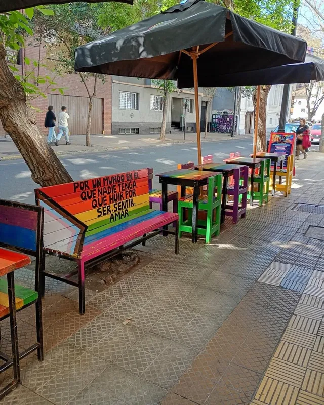 Terraza en la vereda con mesas arcoíris y la banca que dice «Por un mundo en el que nadie tenga que huir por ser, sentir, amar»