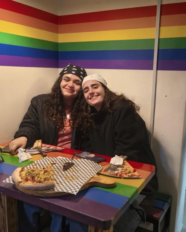 Dos personas abrazadas en una mesa arcoíris de adentro, con una pizza