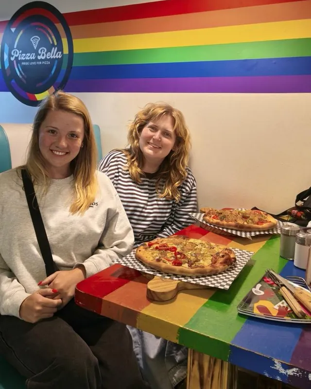 Dos personas con sus pizzas en una mesa arcoíris, junto al logo de Pizzabella