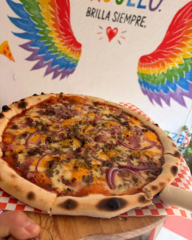 Pizza Lela frente al muro de alas arcoíris que dice «Orgullo. Brilla siempre.»